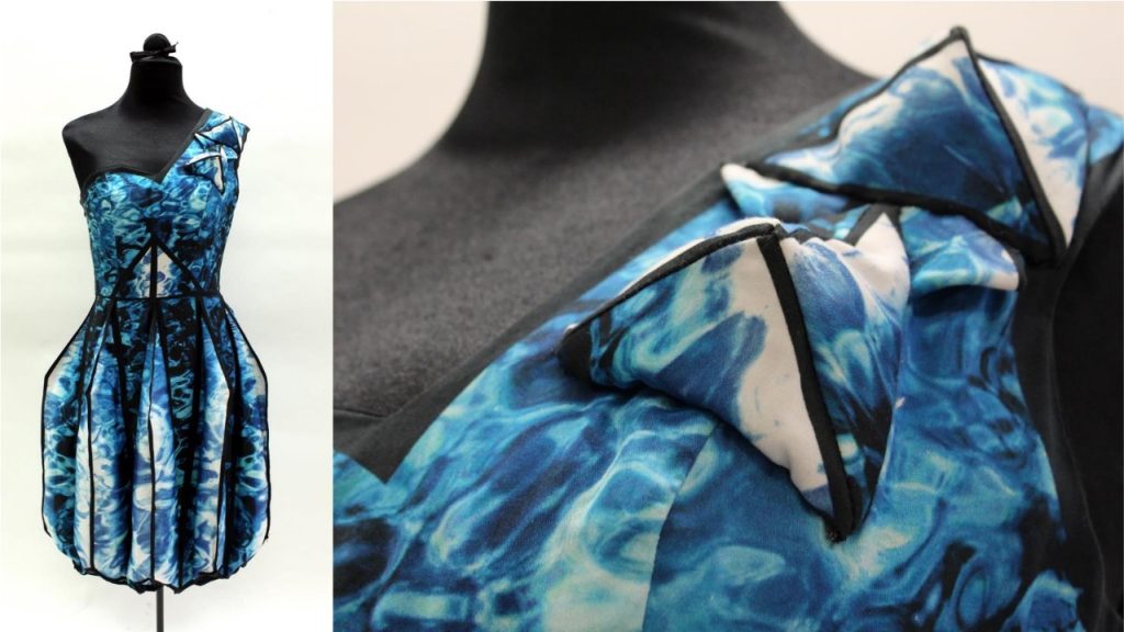 Alt text: "A mannequin displays a dress with a vibrant blue, water-like pattern. The one-shoulder design features bold geometric shapes and flowing fabric."