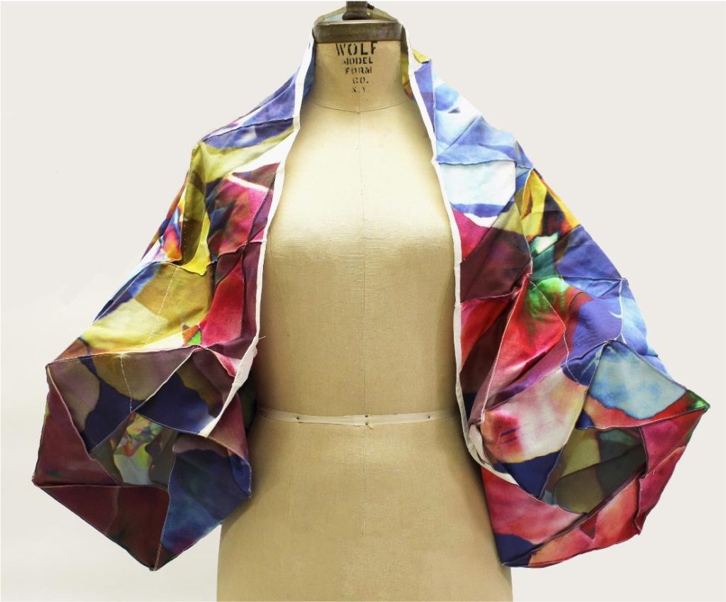 A mannequin displays a vibrant, multicolored artistic shawl draped over its shoulders. The fabric is rich with abstract patterns and bold hues, creating a playful, creative vibe.